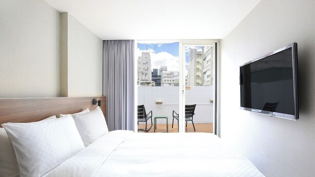 Een hotelkamer met een bed, een televisie, een balkon met stoelen en uitzicht op de stad.