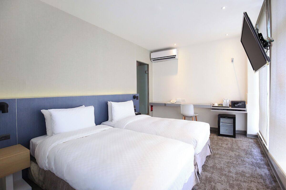 Ein Hotelzimmer mit zwei Betten, einem Schreibtisch, einem Sessel, einer Klimaanlage und einem an der Wand befestigten Fernseher.