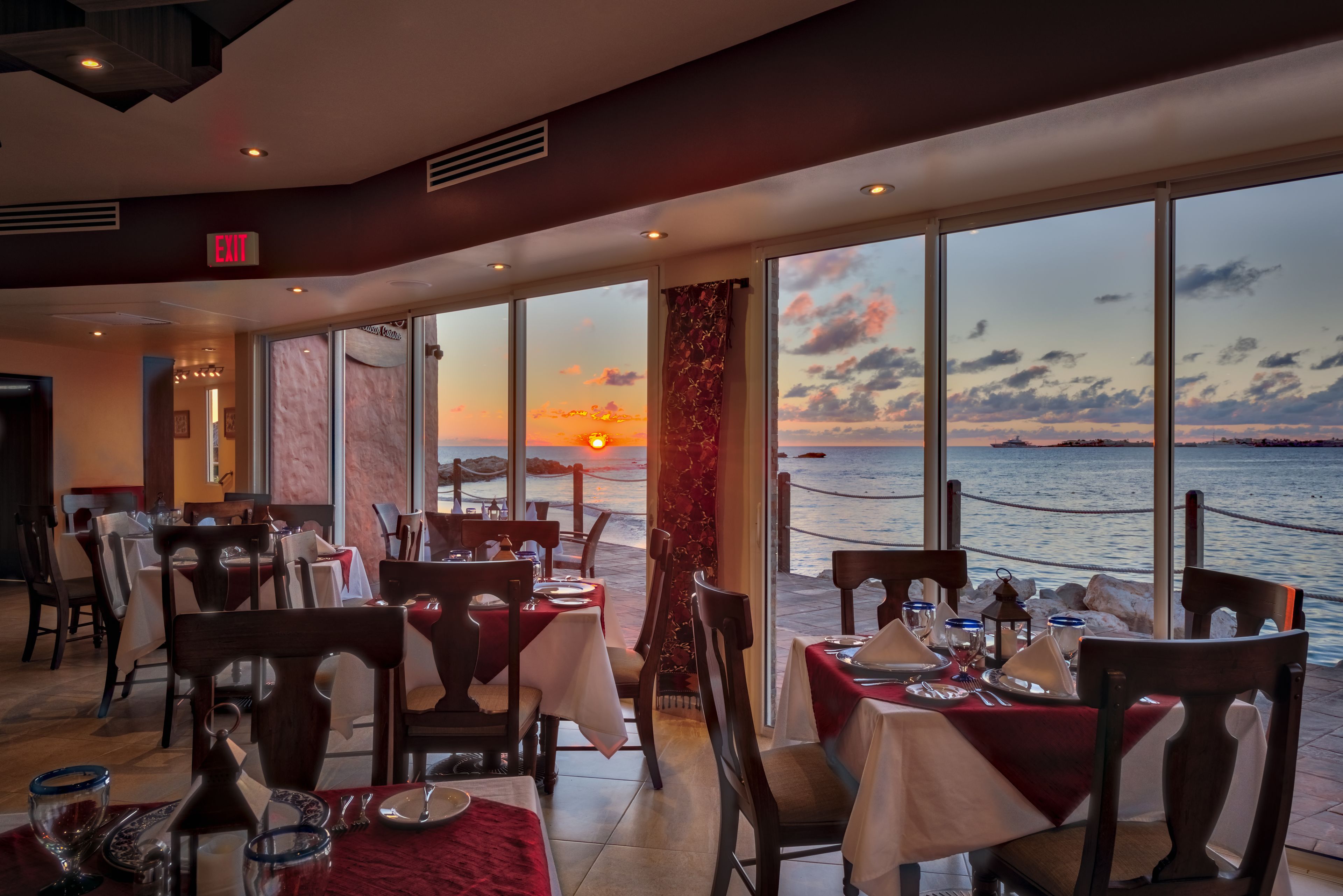 Un restaurante con mesas preparadas para comer, amplios ventanales que ofrecen vista al atardecer sobre el mar, y un letrero de salida sobre la puerta.