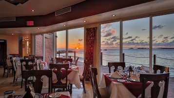 Un restaurante con mesas preparadas para comer, amplios ventanales que ofrecen vista al atardecer sobre el mar, y un letrero de salida sobre la puerta.