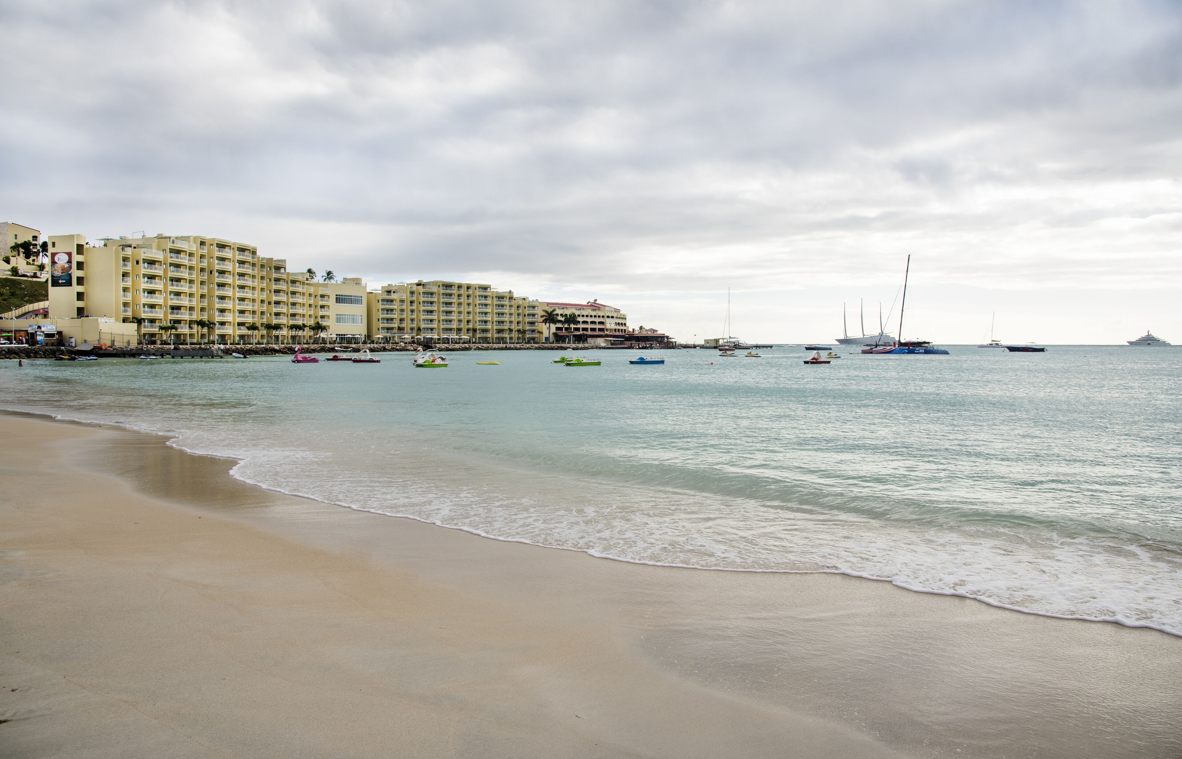 Ein Strand mit einem Hotel im Hintergrund, ruhigem Wasser und Booten.