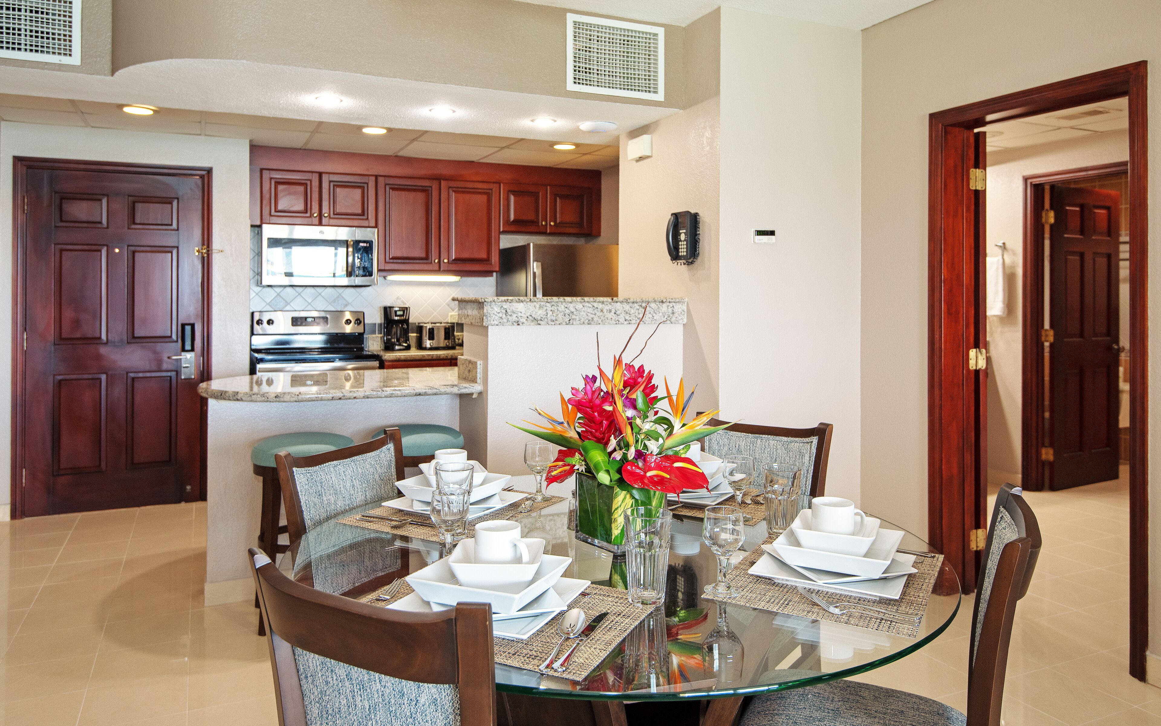 A dining area with a glass table, chairs, and a centerpiece. There is a kitchen in the background with wooden cabinets and appliances.