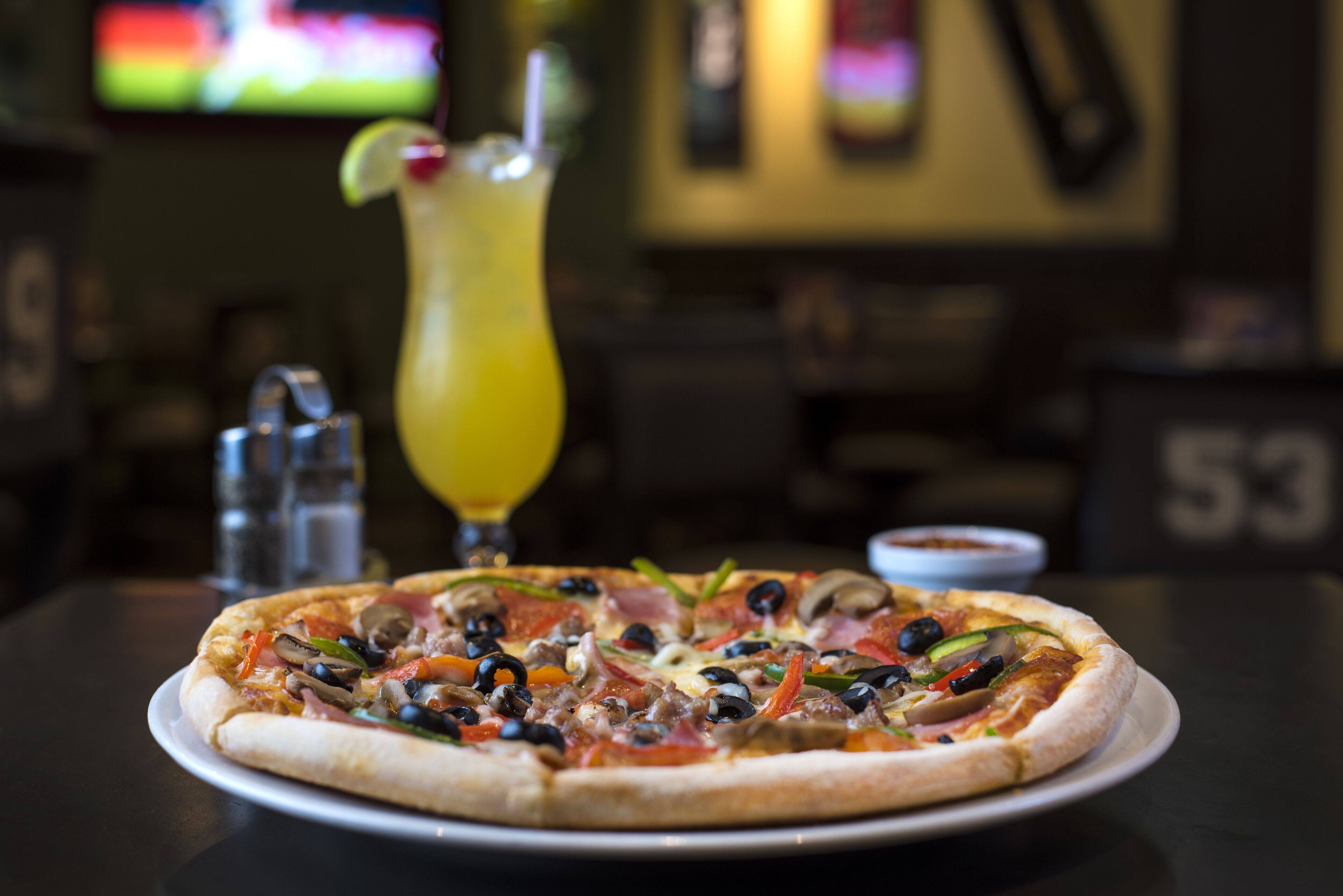 Pizza con diversos ingredientes sobre un plato blanco, un vaso de bebida amarilla con pajita, y saleros y pimenteros sobre una mesa.
