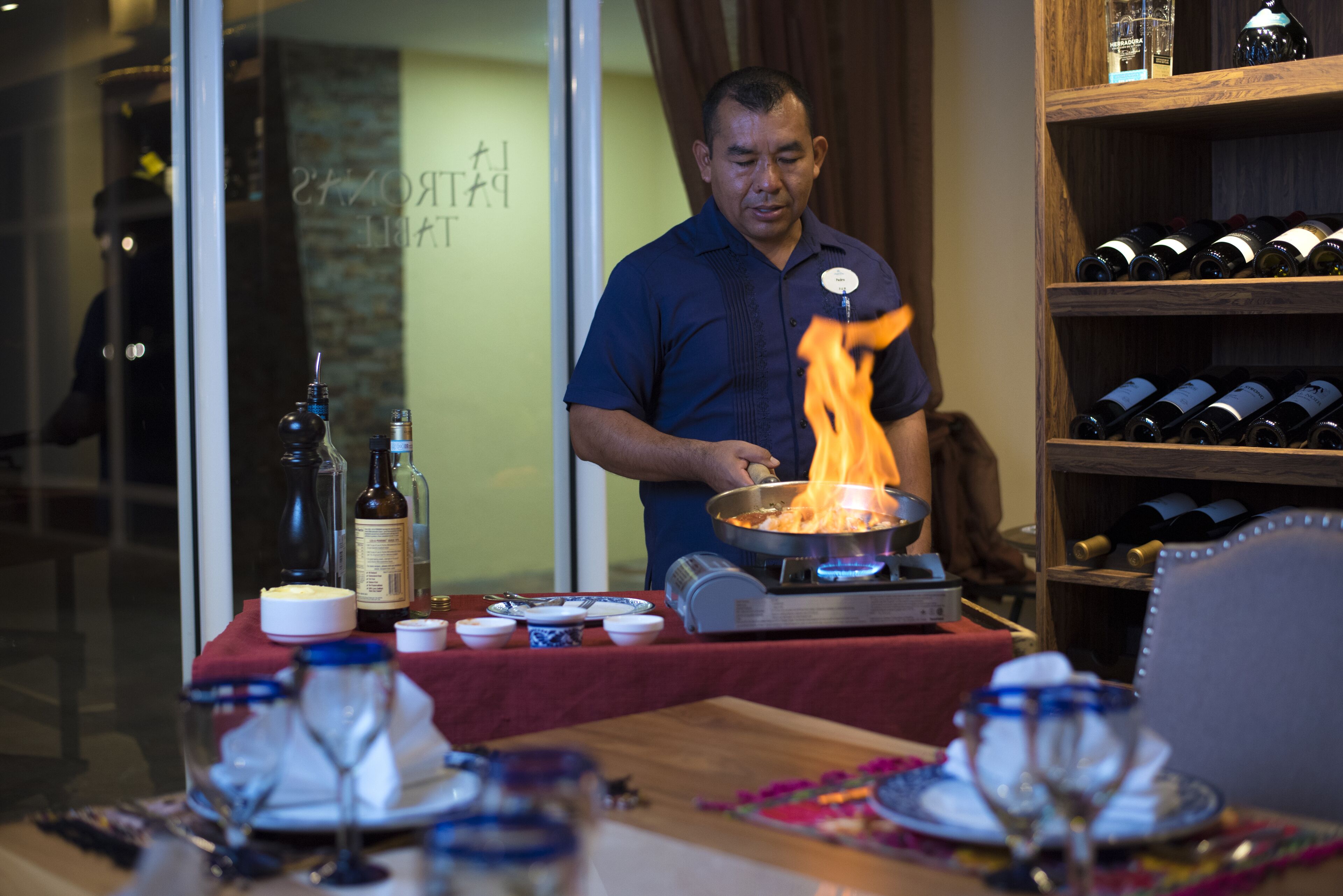 Un comedor con una mesa dispuesta para dos personas, una estantería para vinos al fondo, y alguien cocinando con una sartén sobre una hornilla portátil.