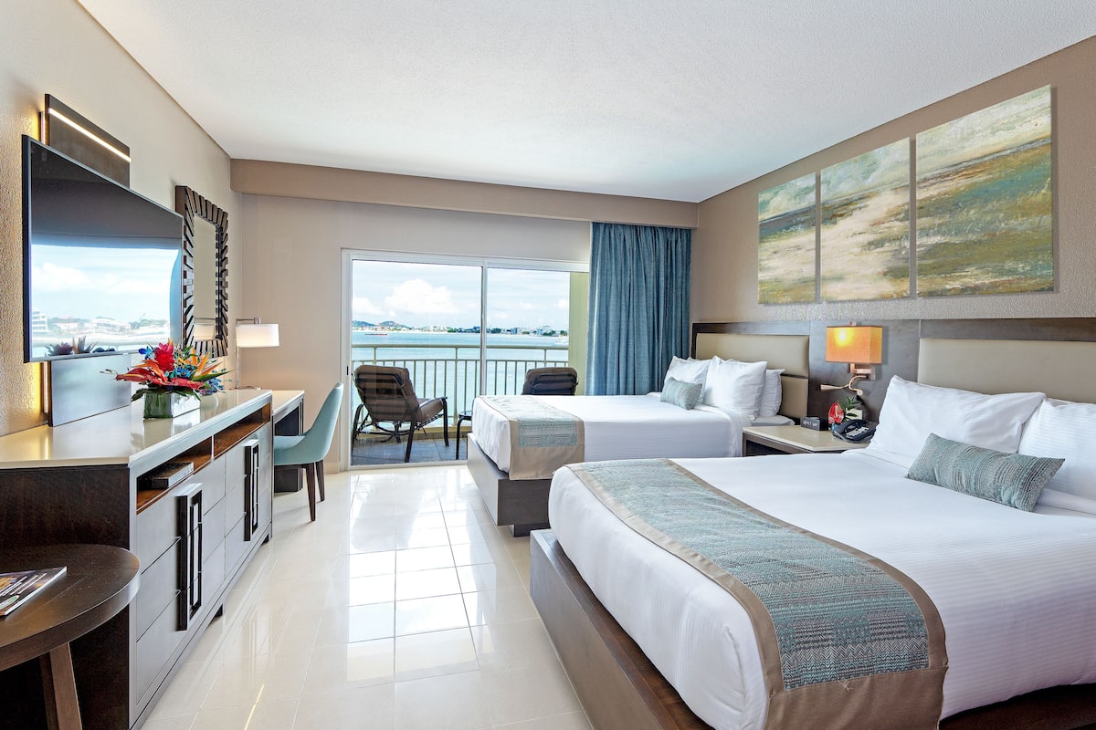 A hotel room with two beds, a desk, a chair, a TV, and a large window with a view of the sea.