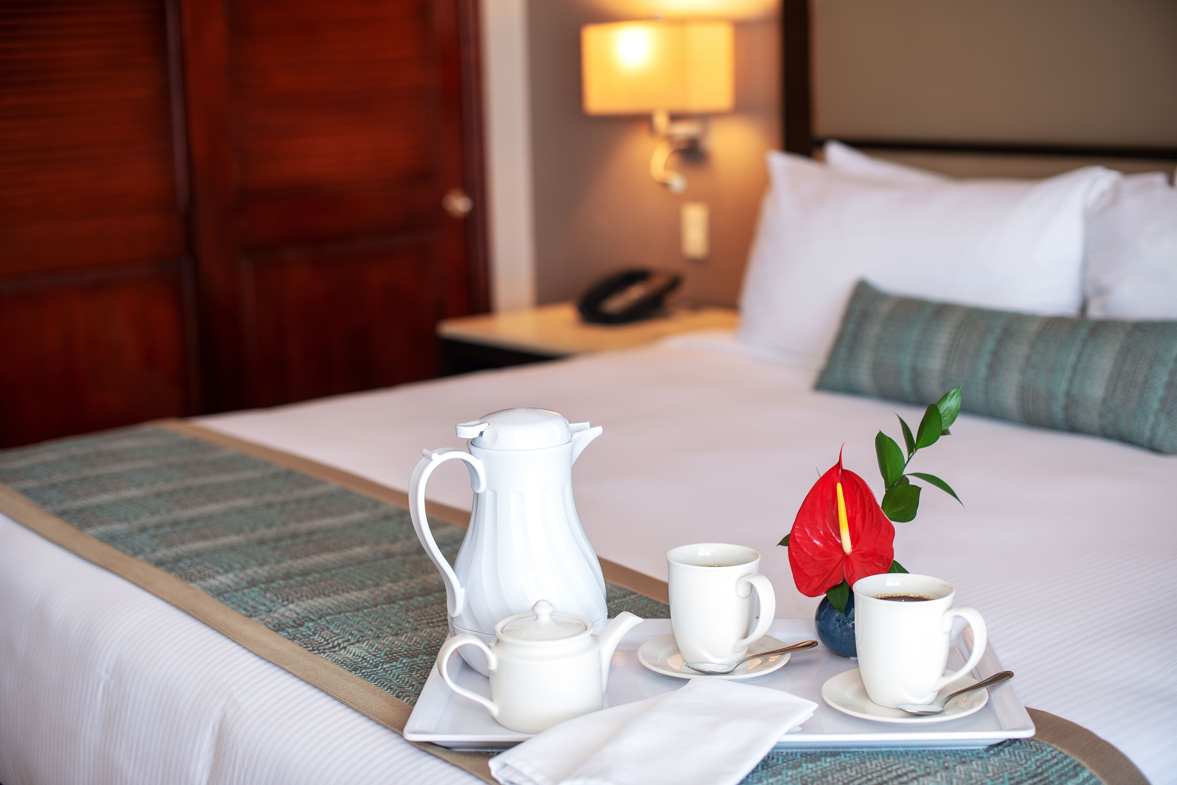 A neatly made bed with a white comforter, a wooden headboard, a bedside table with a lamp, and a tray with a coffee pot, two cups, and a flower.