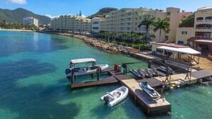 Un hotel frente al mar con un puerto deportivo, aguas turquesas cristalinas y montañas verdes al fondo.