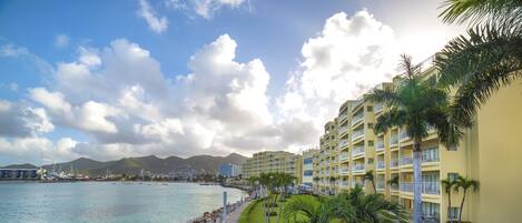 A coastal hotel with a paved walkway along the water, palm trees, and a view of the sea and mountains in the background.