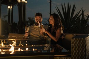 Una pareja brindando con bebidas en una mesa al aire libre, con una chimenea rodeada de mobiliario de mimbre y plantas de palma.