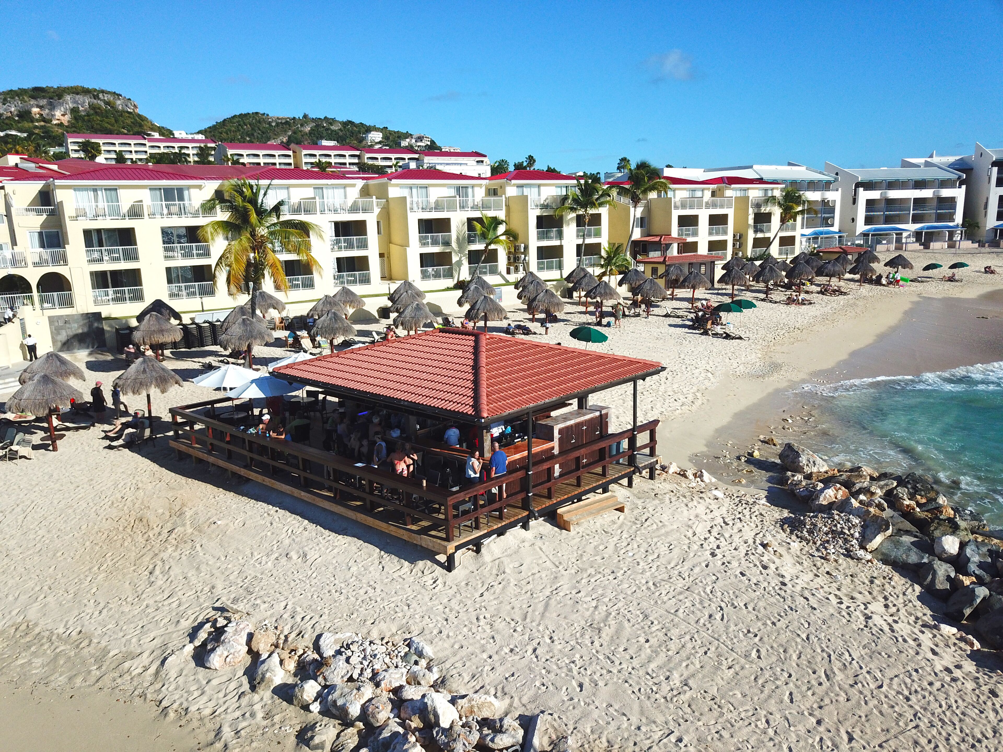Un complexe hôtelier en bord de mer, composé de bâtiments à plusieurs étages, d’un bar de plage et doté de parasols en chaume.