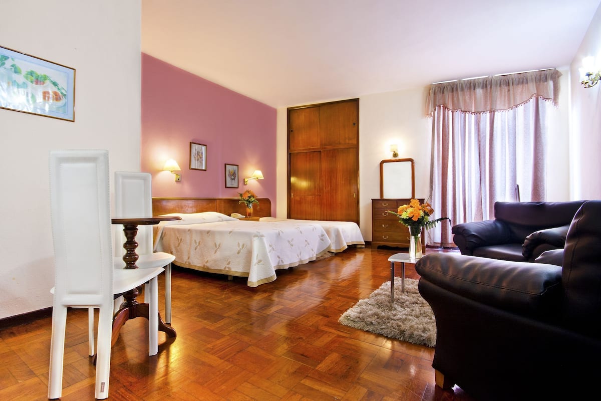 A hotel room with two beds, a wooden floor, a desk, a chair, a sofa, and a window with curtains.