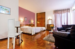 A hotel room with two beds, a wooden floor, a desk, a chair, a sofa, and a window with curtains.