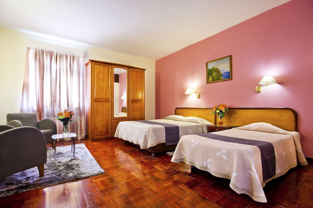 A hotel room with two beds, a wooden wardrobe, a small table with a vase, and a chair.