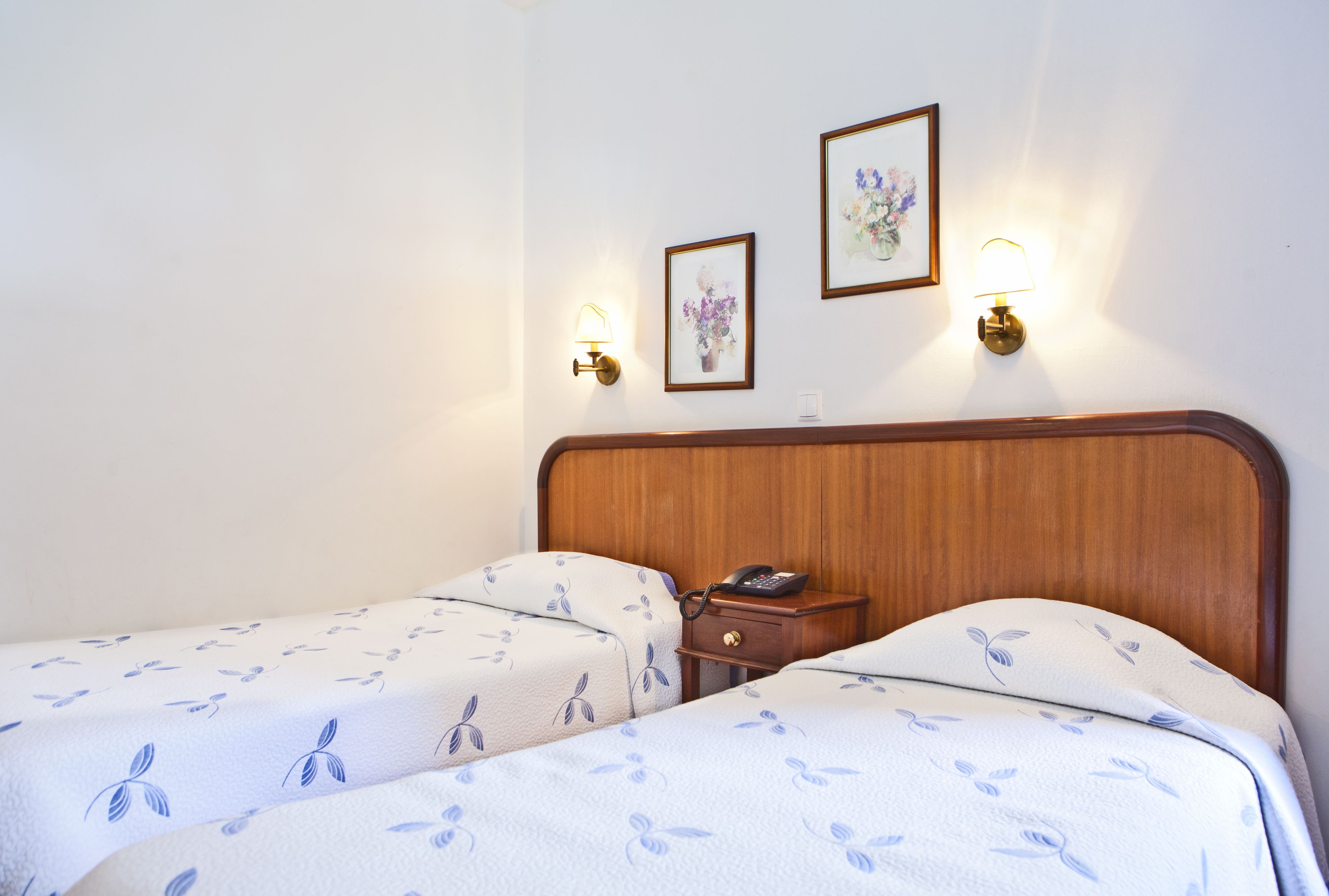 A hotel room with two beds, each with a floral-patterned bedspread, a wooden headboard, bedside tables with lamps, and framed artwork on the wall.