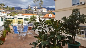 A terrace with chairs and potted plants, overlooking a town with buildings and a mountain in the background. - Residencial Colombo (Funchal)