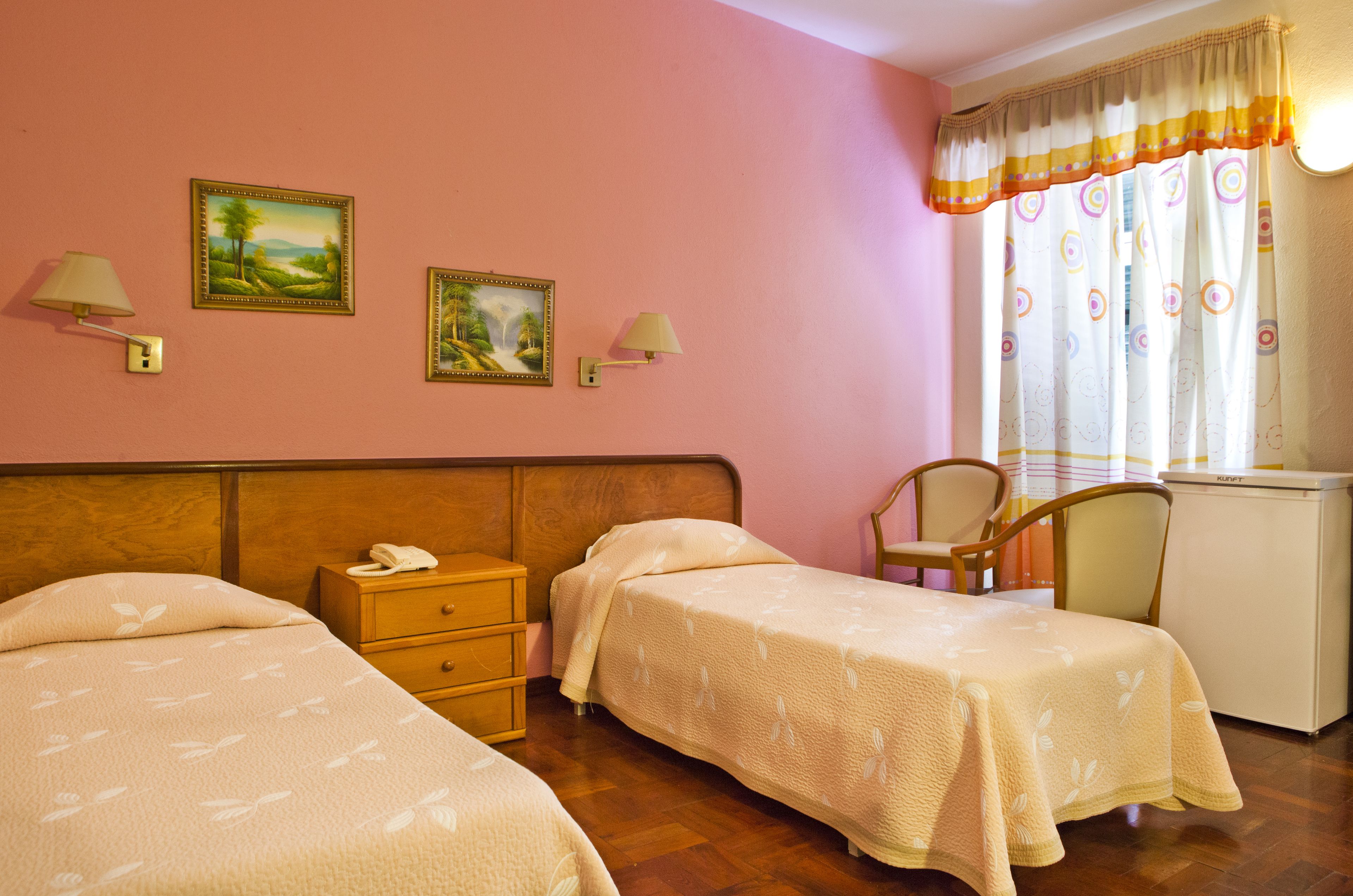 Une chambre d’hôtel avec deux lits, une tête de lit en bois, un bureau avec un téléphone, deux lampes fixées au mur et une fenêtre avec des rideaux.