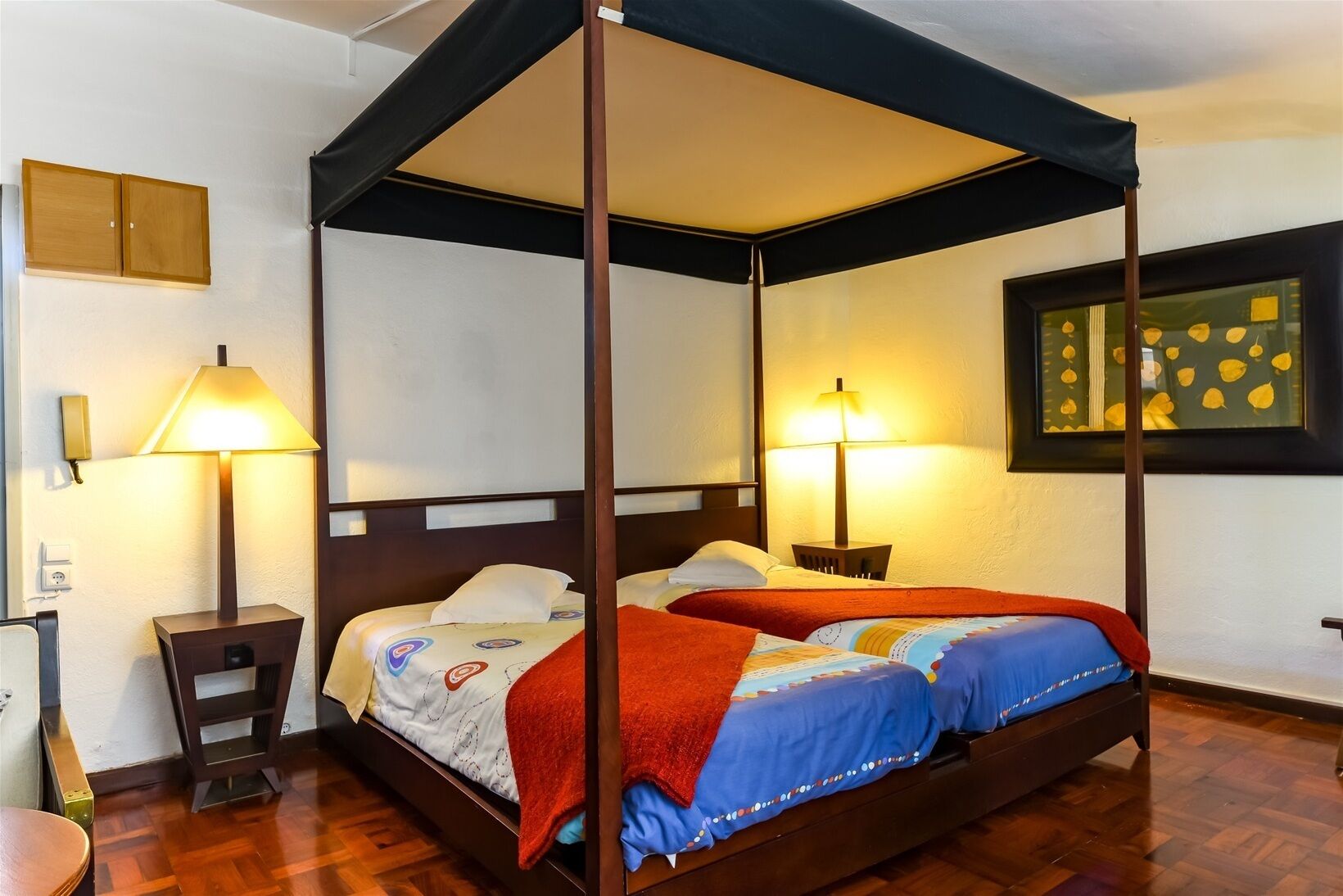 Un lit à baldaquin à quatre poteaux, avec deux tables de chevet, chacune équipée d’une lampe, dans une pièce au plancher en bois et ornée d’un tableau encadré au mur.