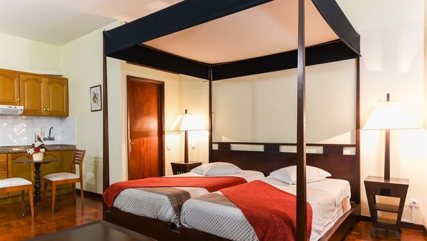 A four-poster canopy bed with two beds, wooden furniture, a small kitchen area, and a dining table. - Residencial Colombo (Funchal)