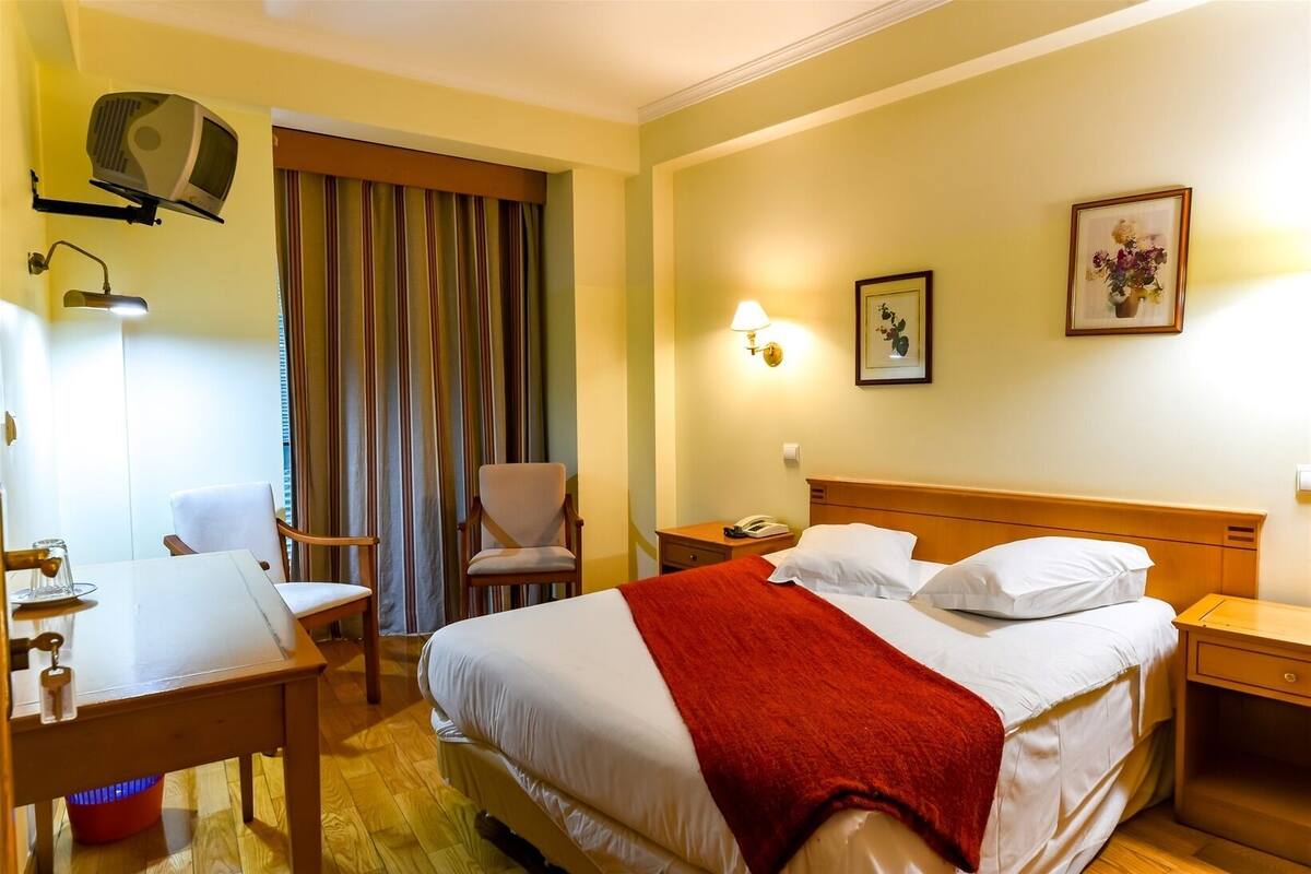 A hotel room with a bed, a desk, a chair, a lamp, a telephone, and a picture on the wall.