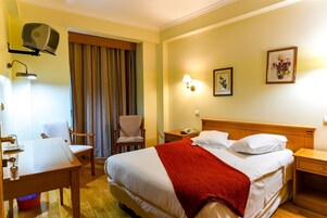 A hotel room with a bed, a desk, a chair, a lamp, a telephone, and a picture on the wall.