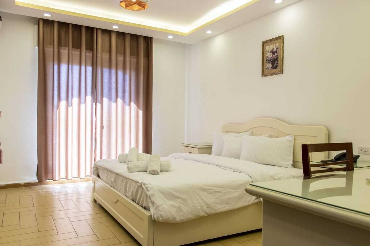 A hotel room with a bed, a bedside table, a chair, a window with curtains, and a painting on the wall.