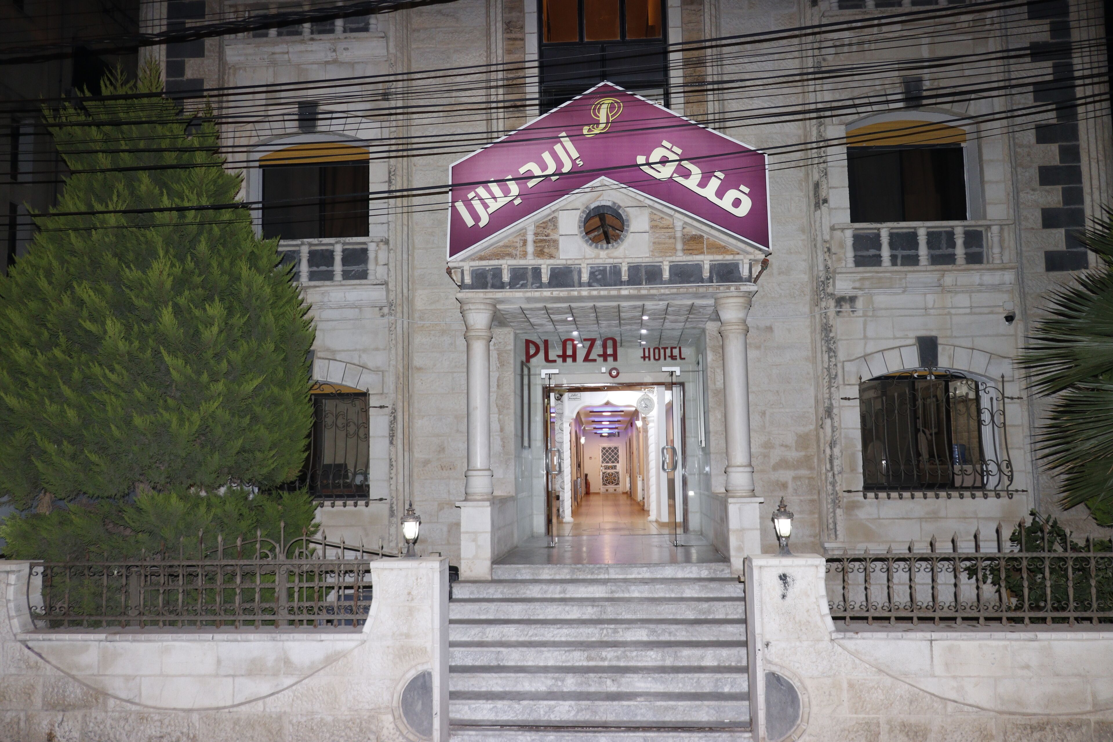 A hotel entrance with a sign in Arabic, a staircase leading up, and a large tree to the left.