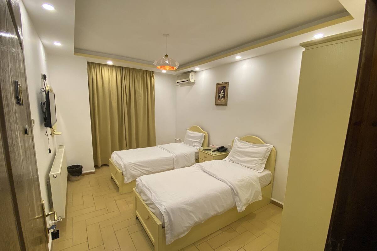 A hotel room with two beds, a ceiling light, an air conditioning unit, a door, and a window with curtains.