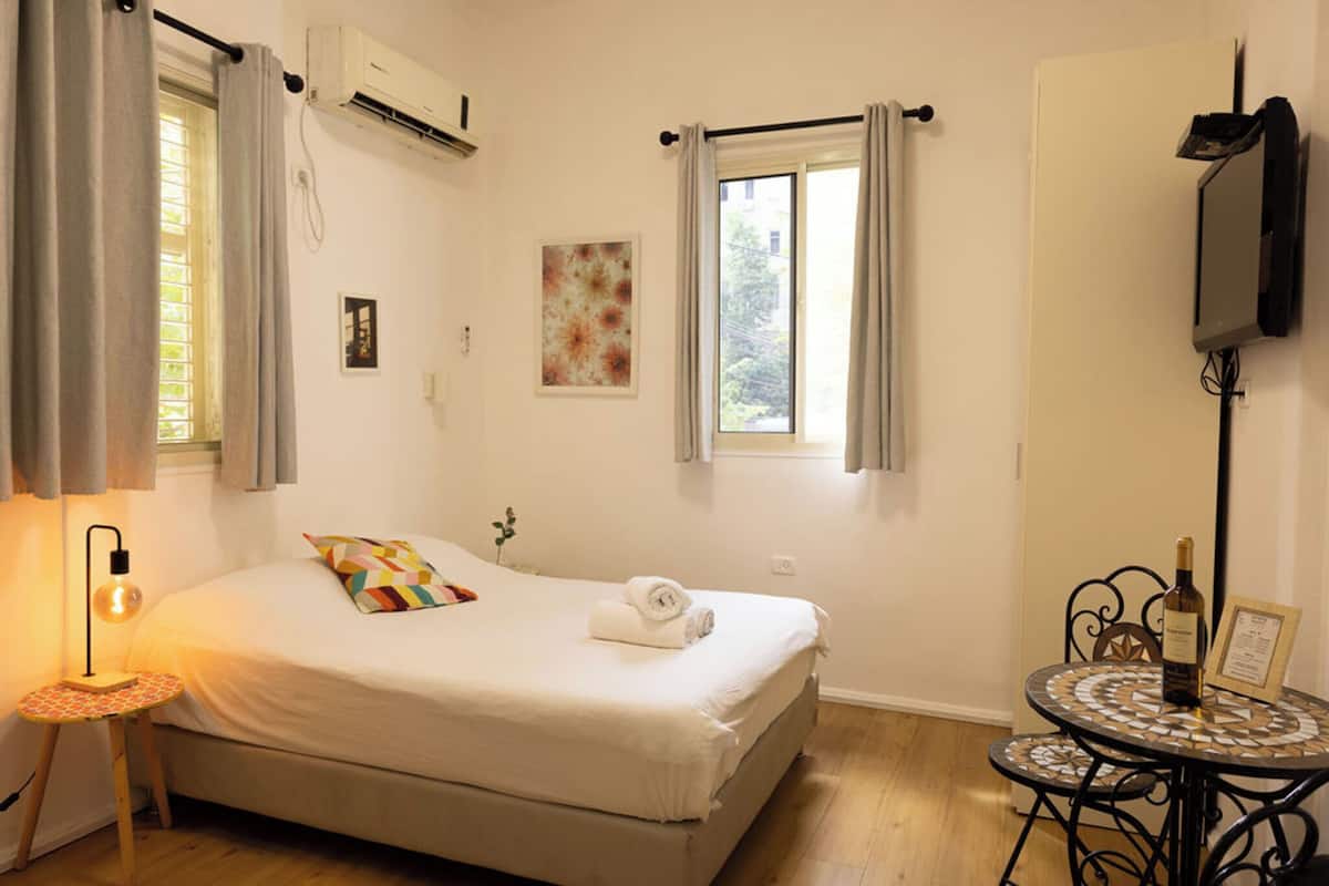 Ein Schlafzimmer mit einem Bett, einem Nachttisch mit Lampe, einem Fenster mit Vorhängen, einem an der Wand montierten Fernseher und einem kleinen Tisch mit einer Flasche und Speisekarte.