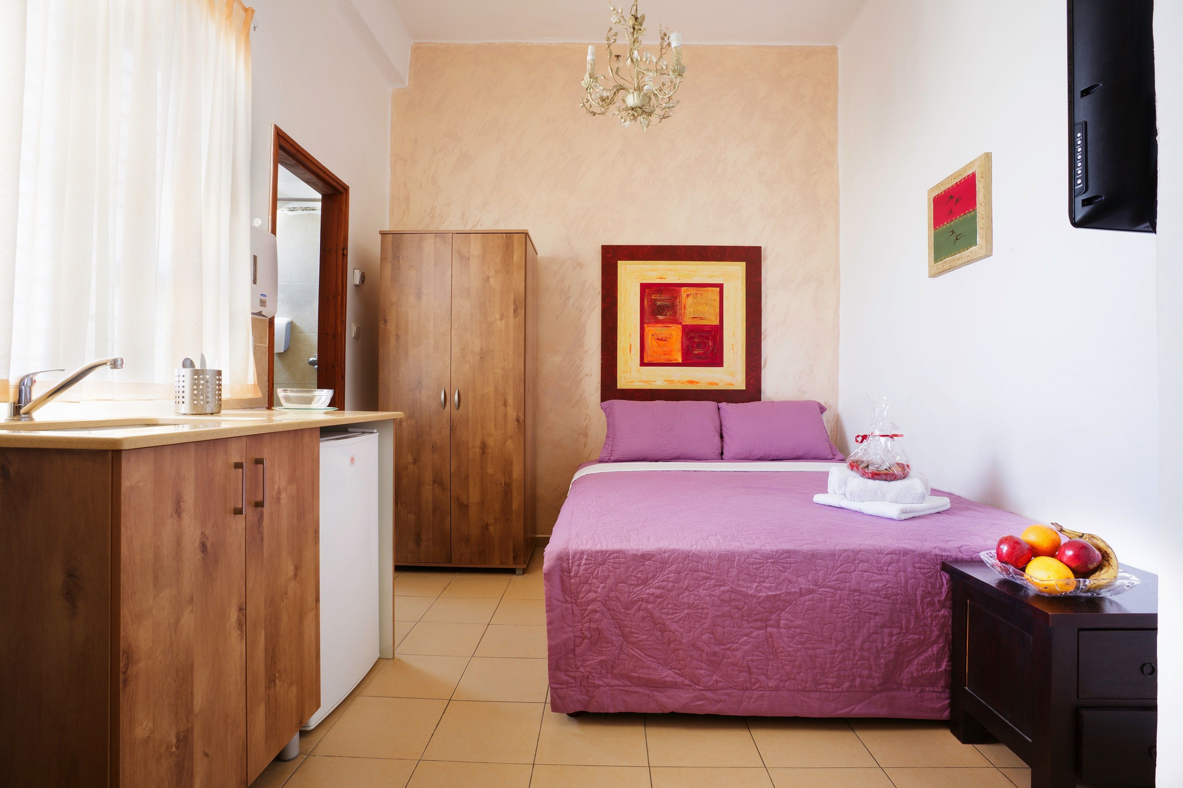 Ein Schlafzimmer mit einem Bett, einem Holzschrank, einer kleinen Küchenzeile mit Spüle und einer Obstschale auf dem Nachttisch.