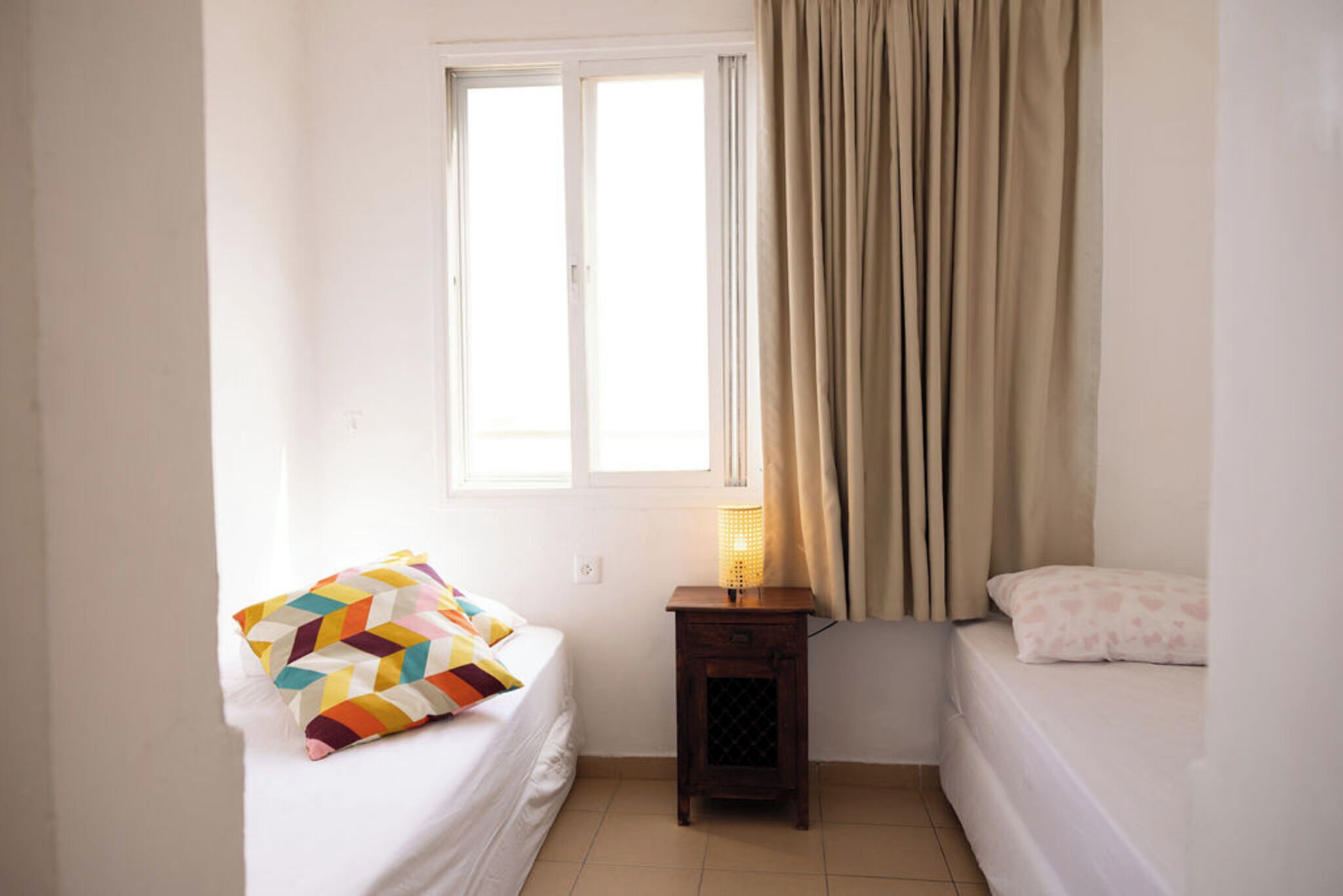 Een kleine, goed verlichte slaapkamer met een enkel bed, een nachtkastje, een lamp en een raam met gordijnen.