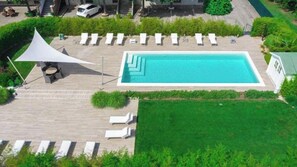 Una piscina rectangular con un deck blanco alrededor, acompañada de sillones de descanso y una zona con sombra.