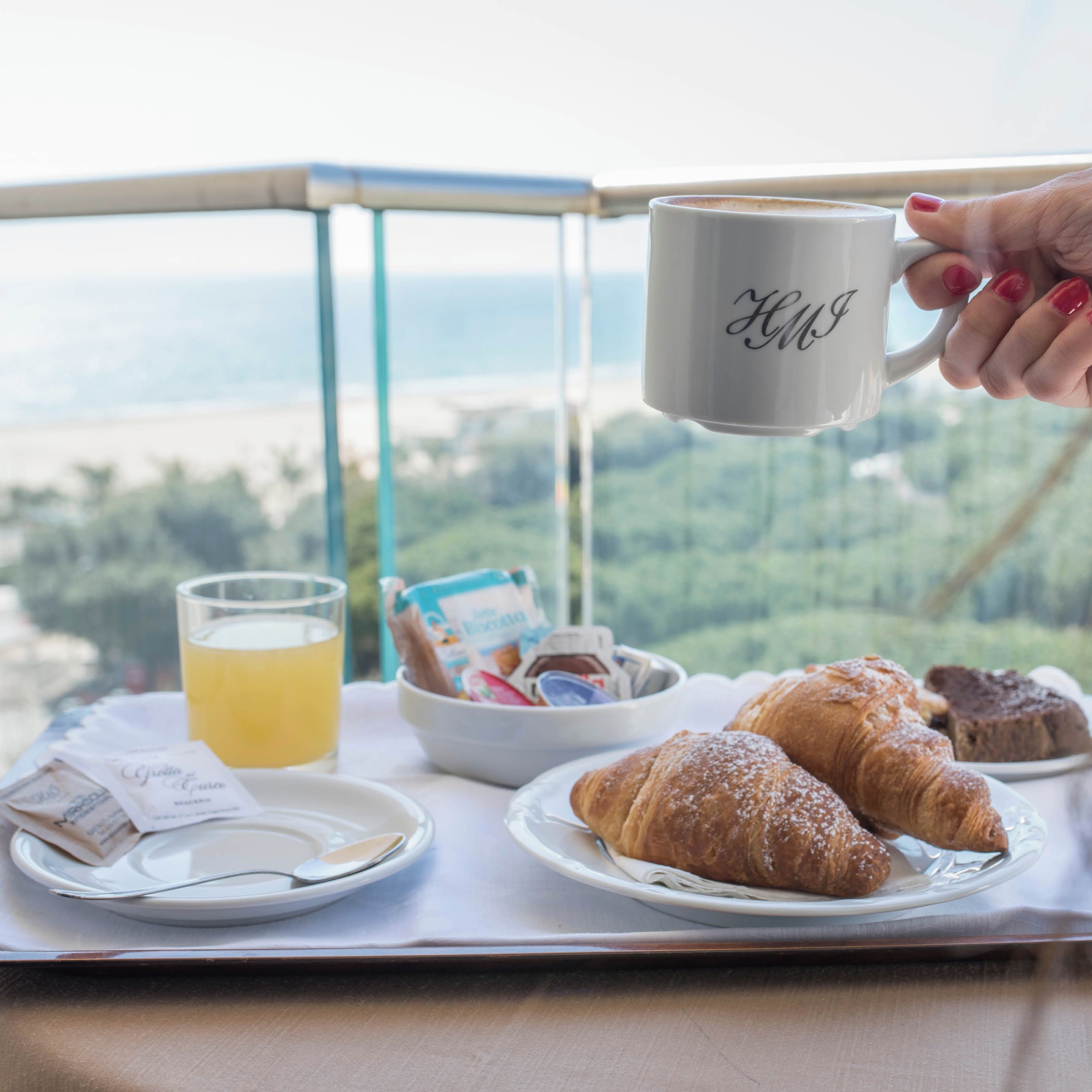 Un vassoio colazione con croissant, un bicchiere di succo d'arancia e una tazza con iniziali, appoggiato su un balcone con vista panoramica.