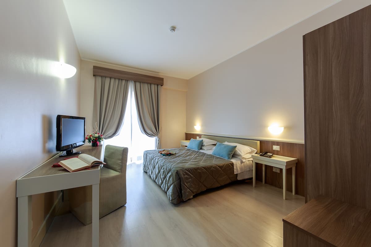 A modern hotel room with a bed, bedside tables, a desk with a computer, and a window with curtains.