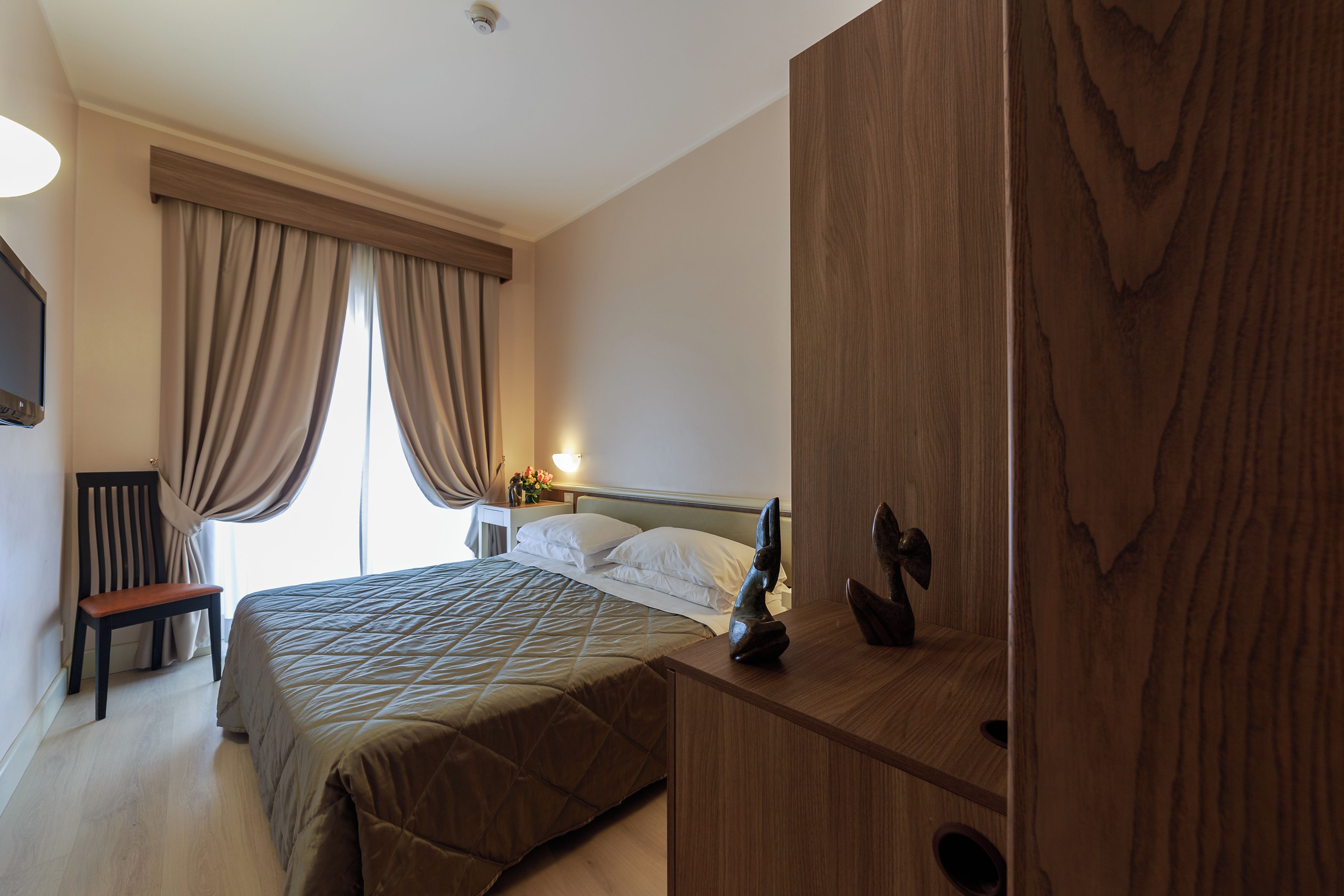 Habitación de hotel con cama, mesita de noche de madera, una silla y un escritorio con lámpara.