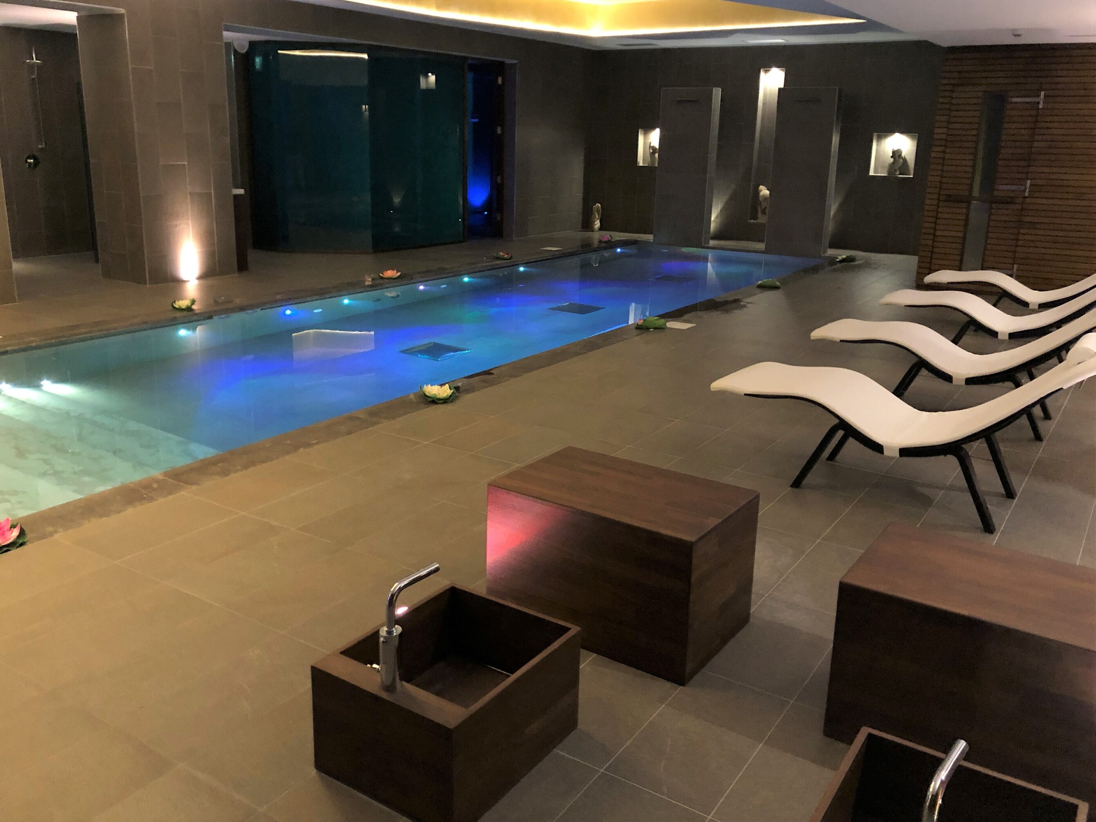Un área de piscina interior moderna con una pileta larga, sillas de descanso y un pequeño espacio de estar con mesas.