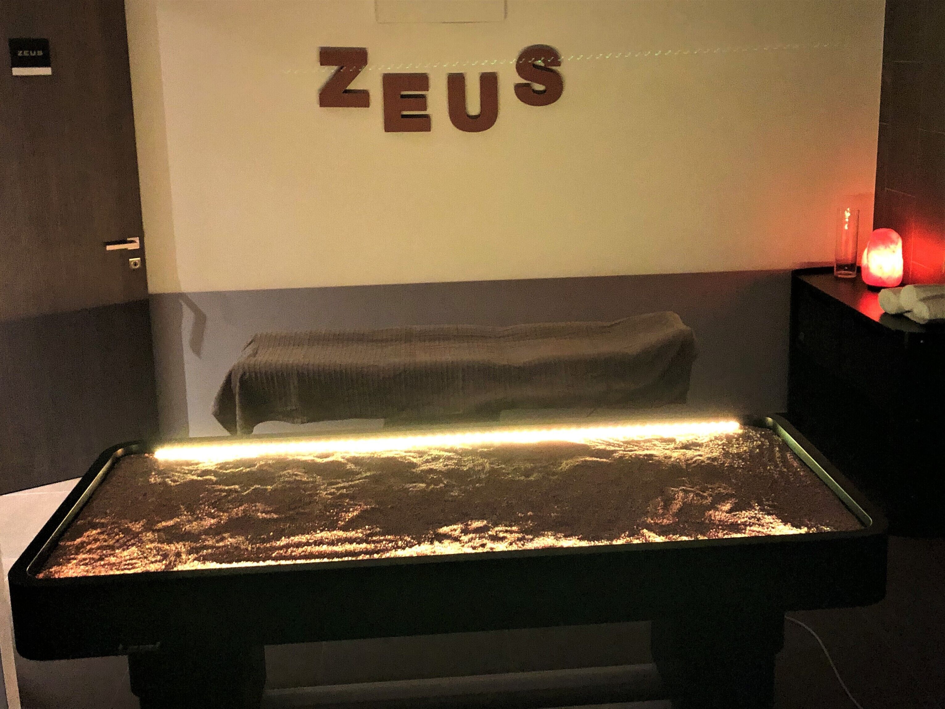 Una stanza con un letto, un cartello appeso al muro con la scritta "ZEUS" e un tavolo con una lampada.