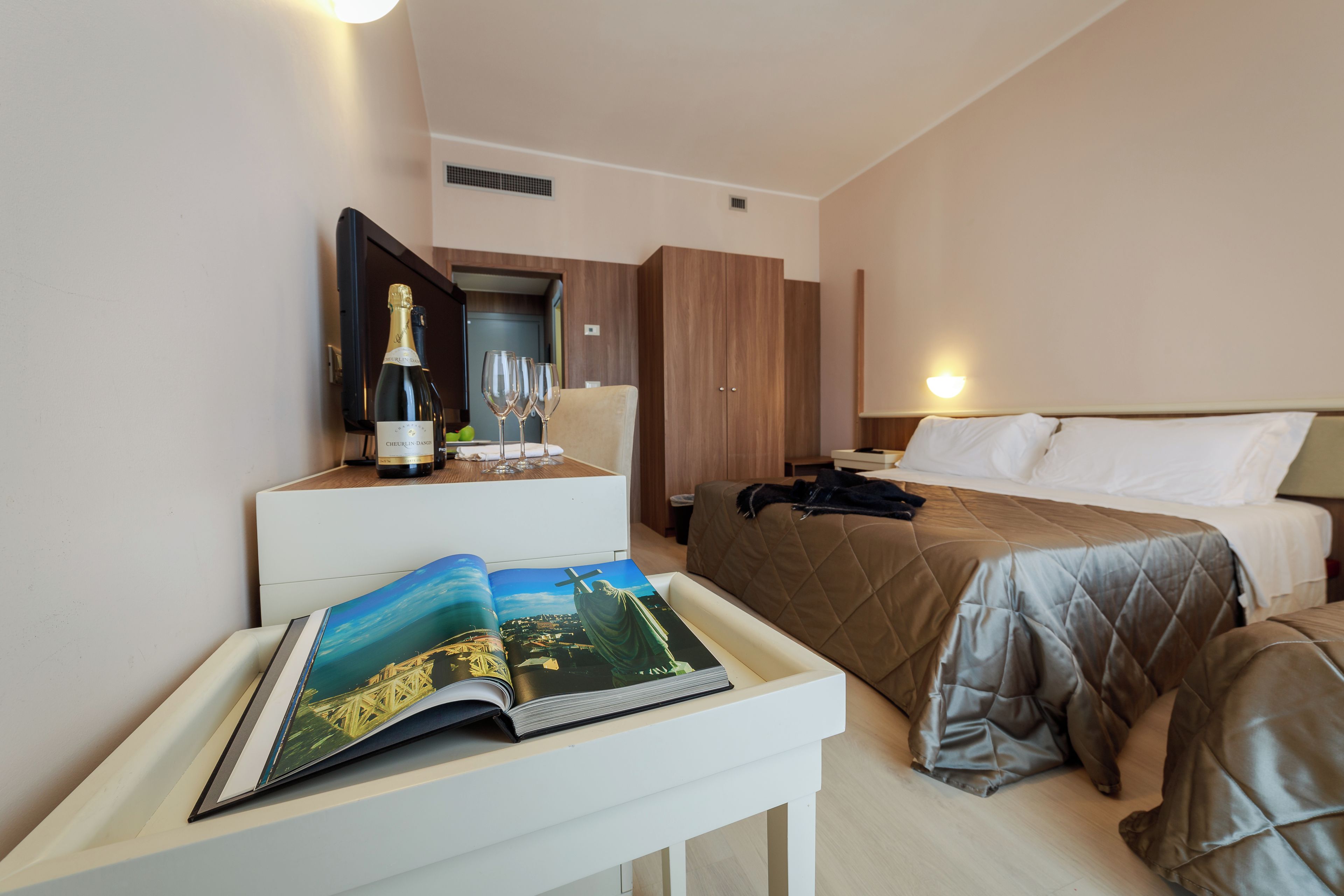 A hotel room with a bed, bedside table, a magazine, a bottle of champagne, and a television.