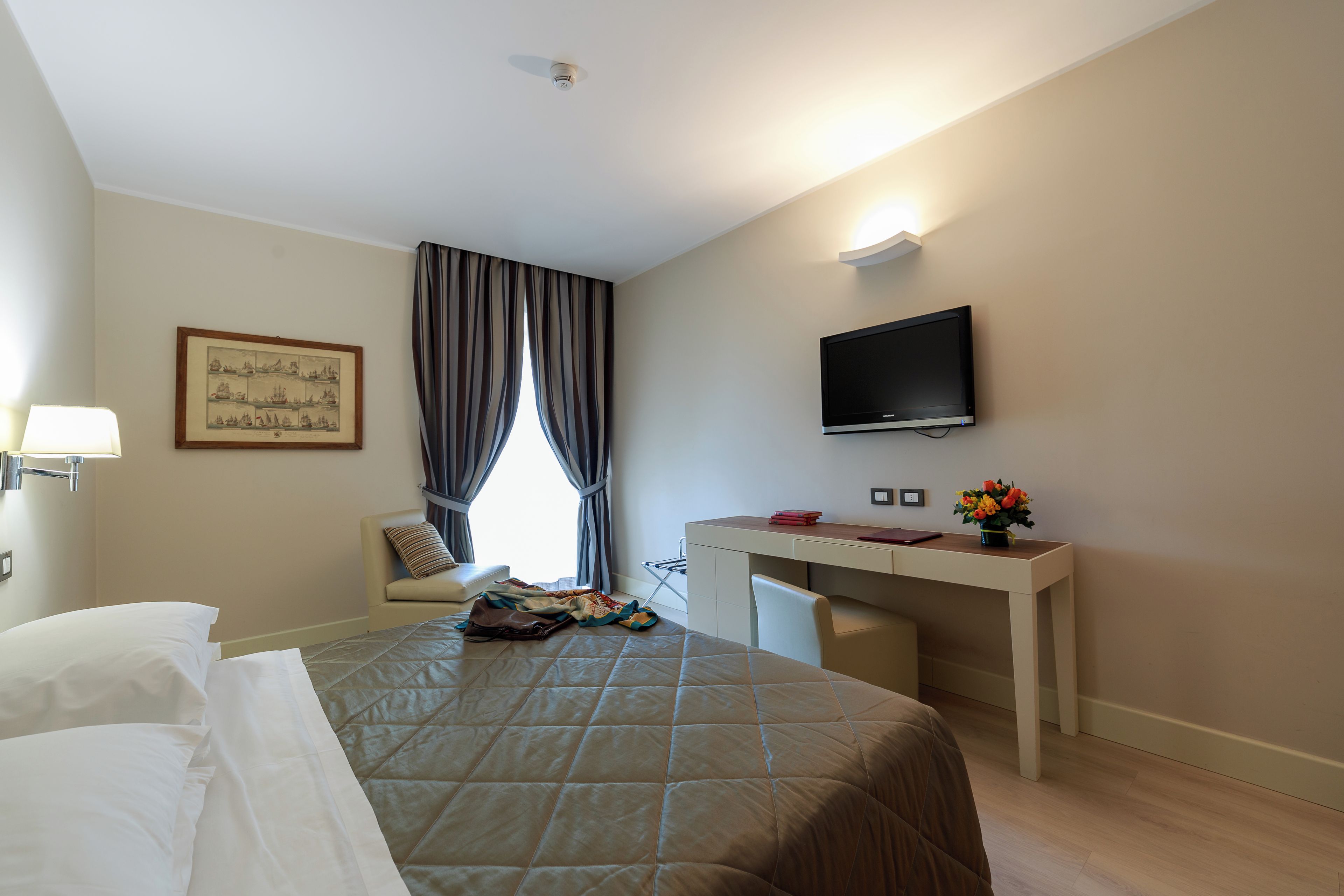 Une chambre d’hôtel comprenant un lit, un bureau, une télévision, une fenêtre avec des rideaux et un tableau accroché au mur.
