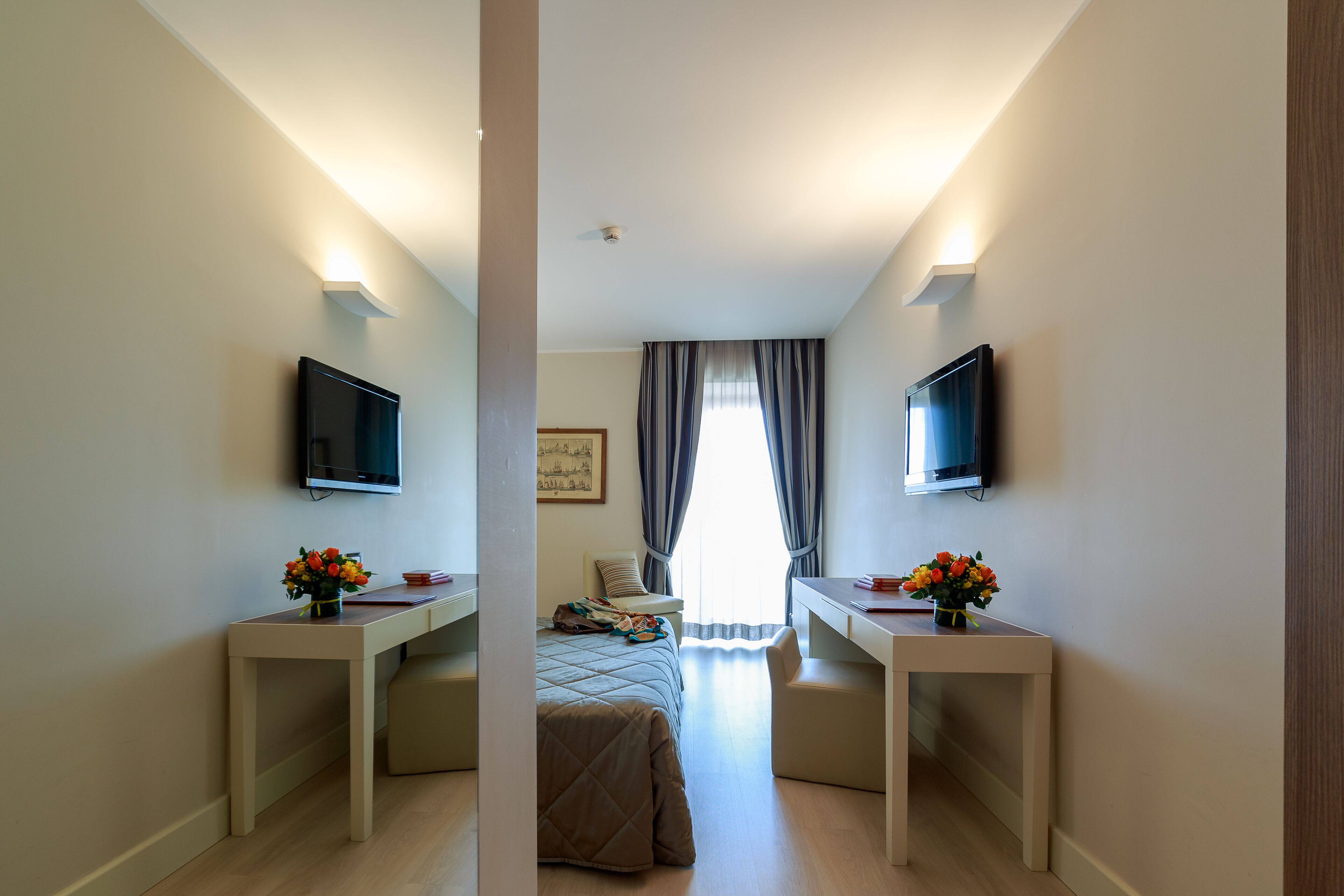 Habitación de hotel con cama, dos mesitas de noche con jarrones de flores, un escritorio, una silla, televisor montado en la pared y una ventana con cortinas.