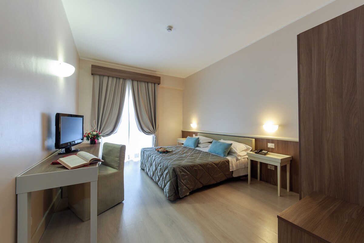 A modern hotel room with a bed, bedside tables, a desk with a computer, and a window with curtains.