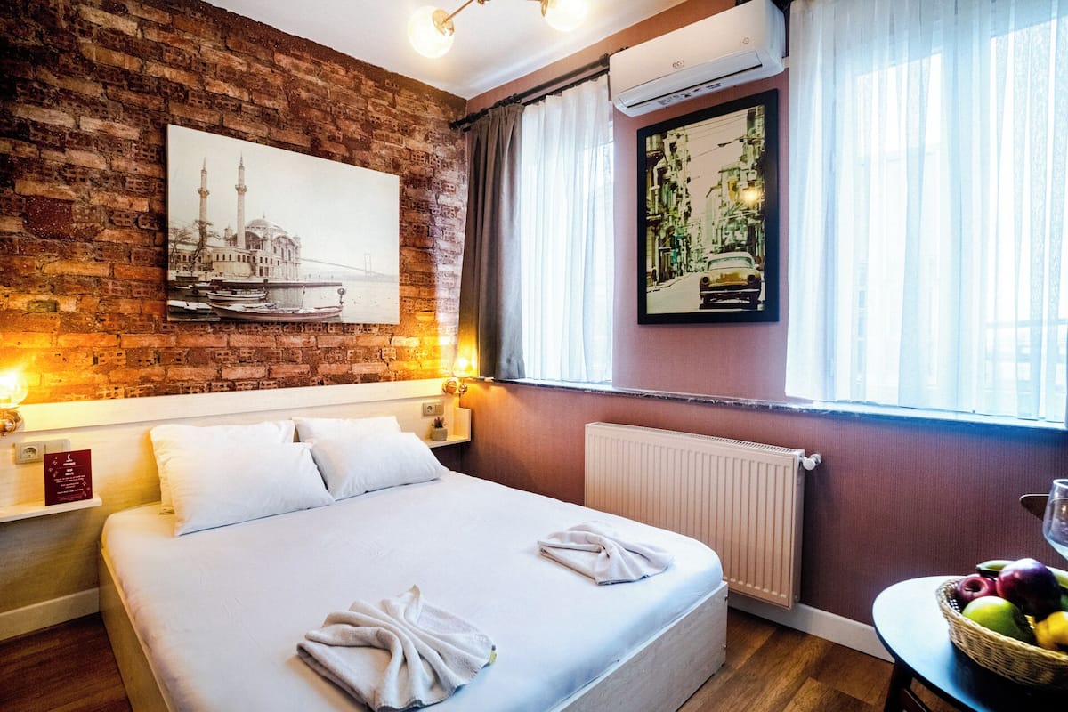 A hotel room with a brick wall, a large bed with two pillows, a framed picture of a cityscape, an air conditioning unit, and a window with curtains.