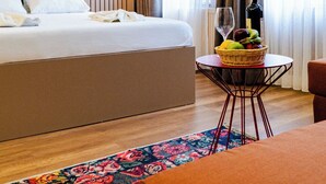 Habitación de hotel moderna con una cama grande, una pequeña mesa de comedor, una cesta de frutas, una botella de vino y una silla roja.