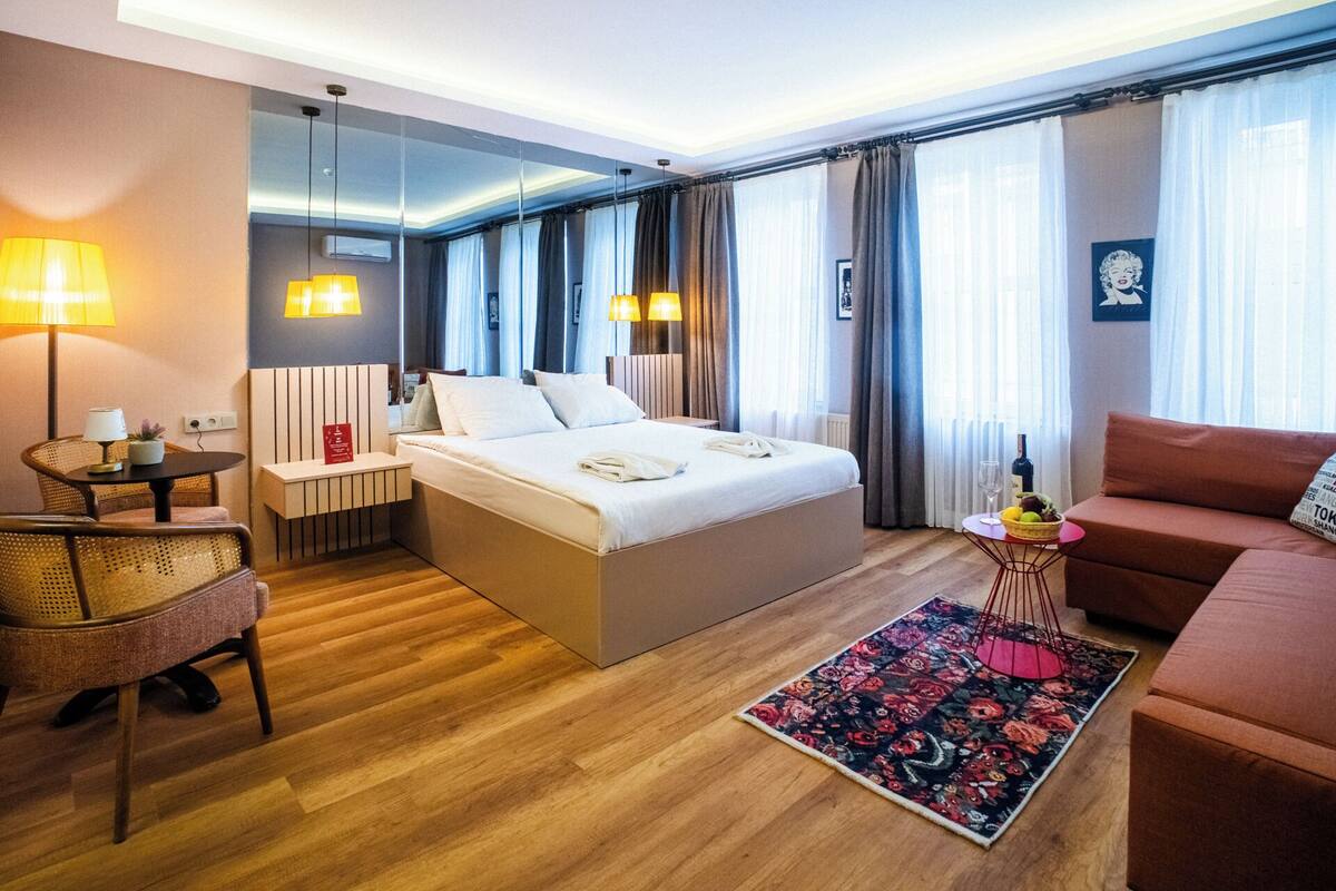 A hotel room with a large bed, a sitting area with a sofa and armchair, a small table with a lamp, and a decorative rug.