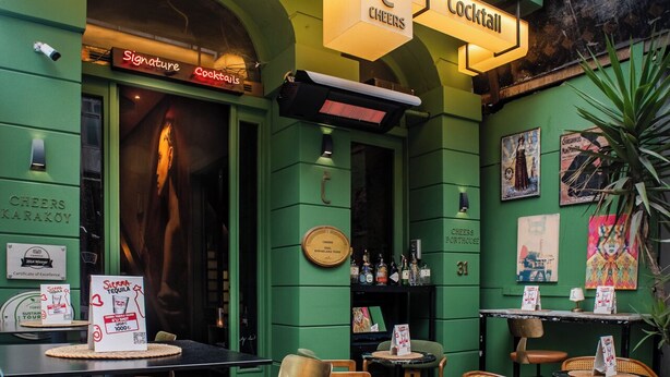 Un área de estar al aire libre en un bar bistró con paredes verdes, un letrero de neón y diferentes opciones de asientos, como taburetes y sillas.