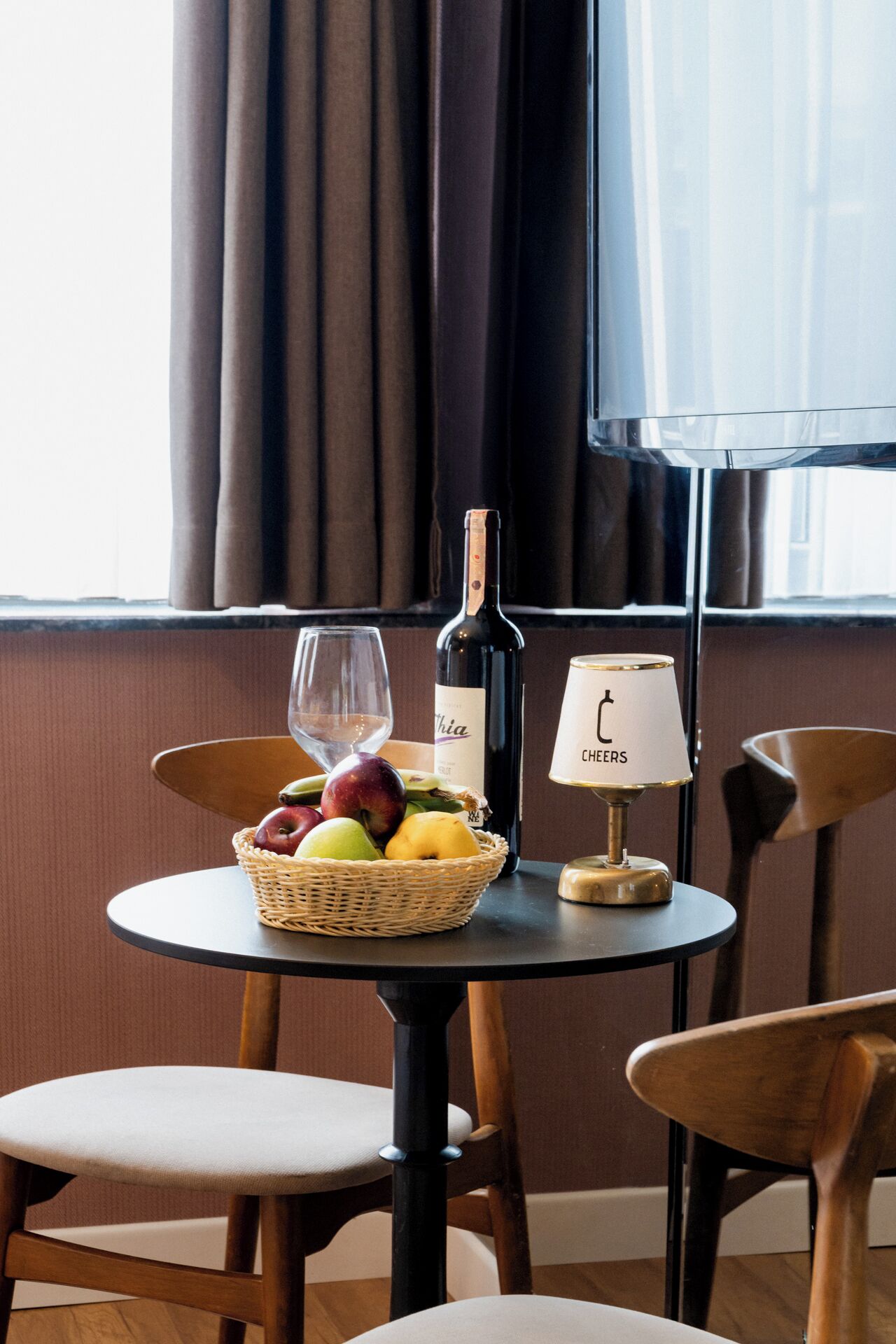 Una mesa con una botella de vino, una copa y una cesta de fruta. Hay una lámpara sobre la mesa y dos sillas alrededor.