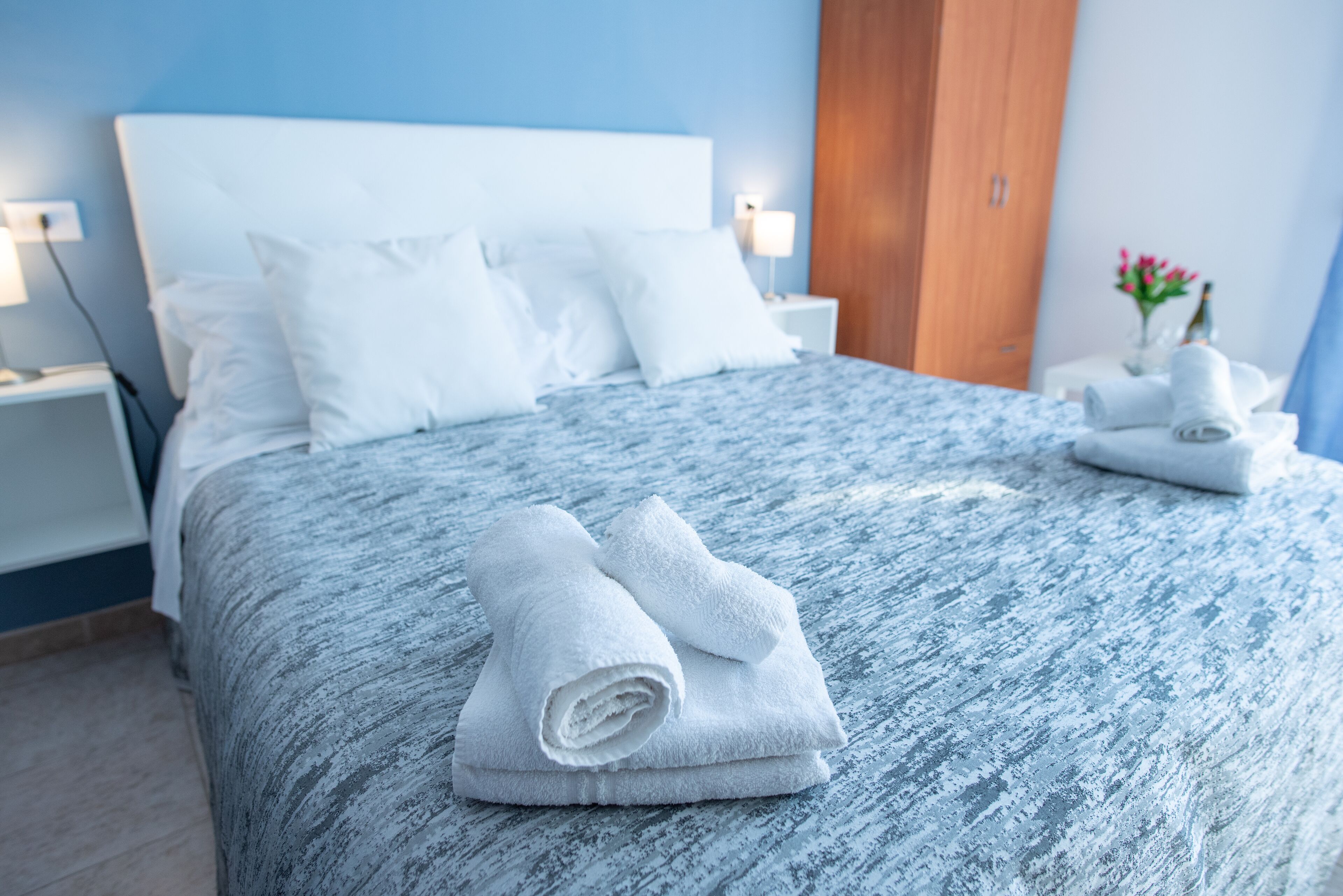 Een net opgemaakt bed met witte lakens, een blauwe geweven sprei, en een houten garderobe op de achtergrond.