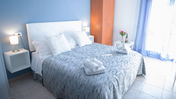 A neatly made bed with white linens, a wooden wardrobe, a bedside table with a lamp, and a window with a blue curtain. - Bed and Breakfast Triscele (Sciacca)