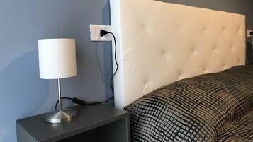 A neatly made bed with a patterned bedspread, a white headboard, and a bedside table with a lamp and charger.