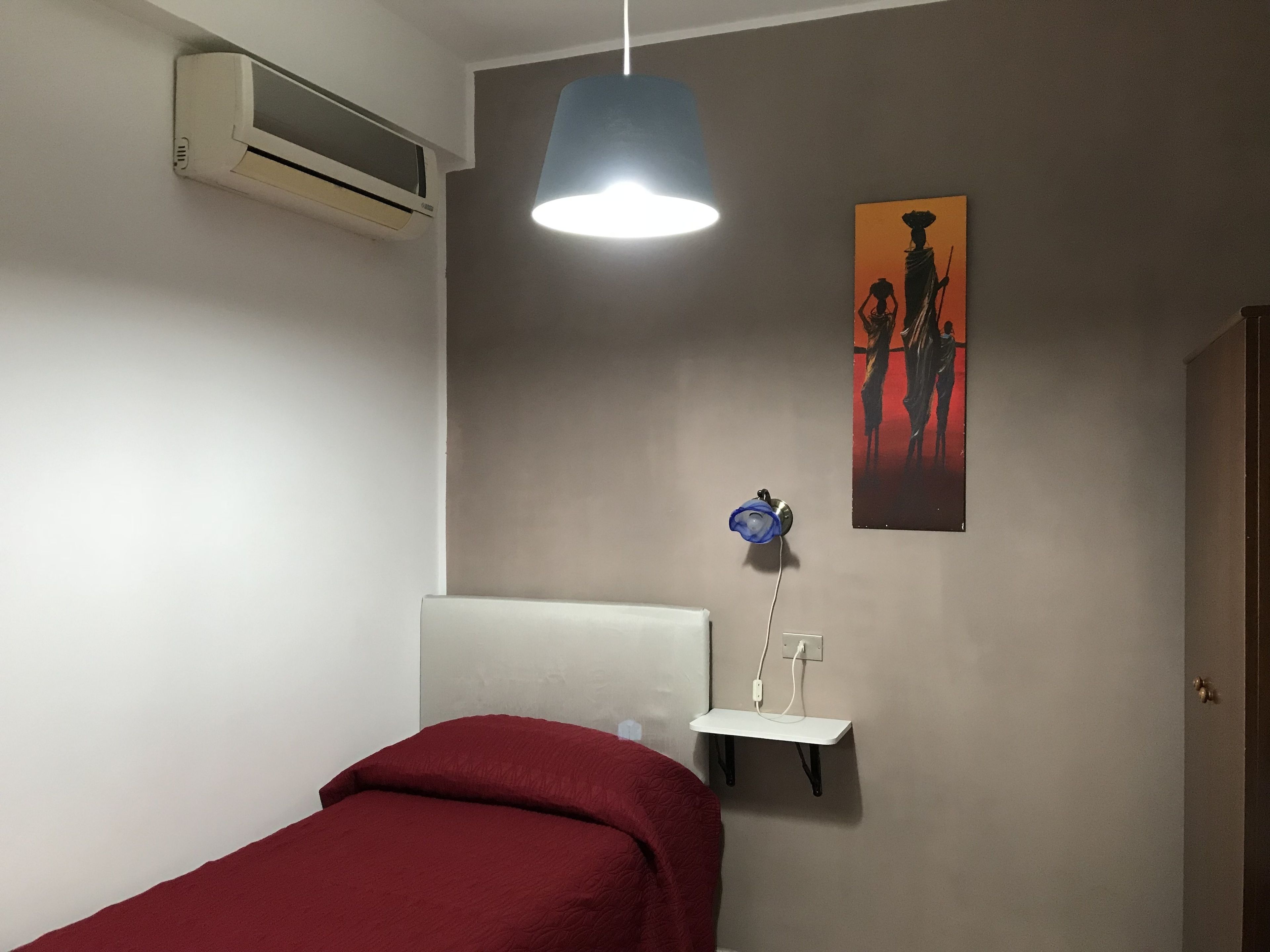 A bedroom with a bed, a wall-mounted air conditioner, a pendant light, a small shelf with a blue vase, and a painting on the wall.