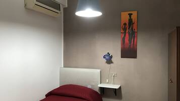 A bedroom with a bed, a wall-mounted air conditioner, a pendant light, a small shelf with a blue vase, and a painting on the wall.